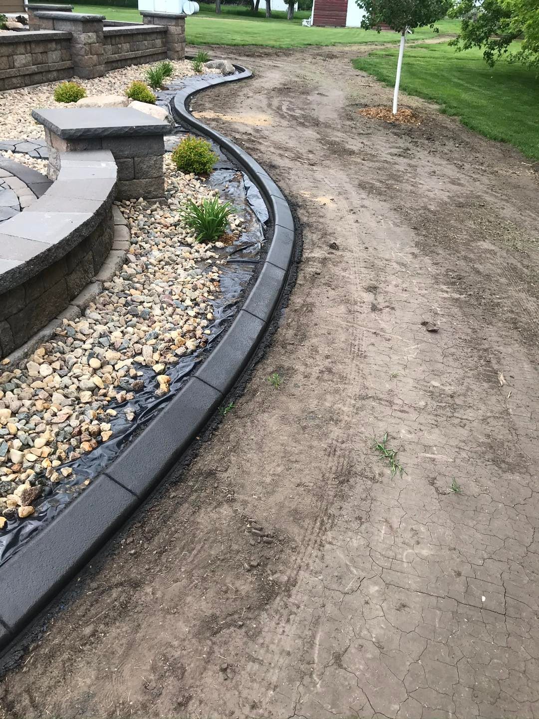 Curved stone border separates gravel landscaping from a dirt path. Green grass in the background.