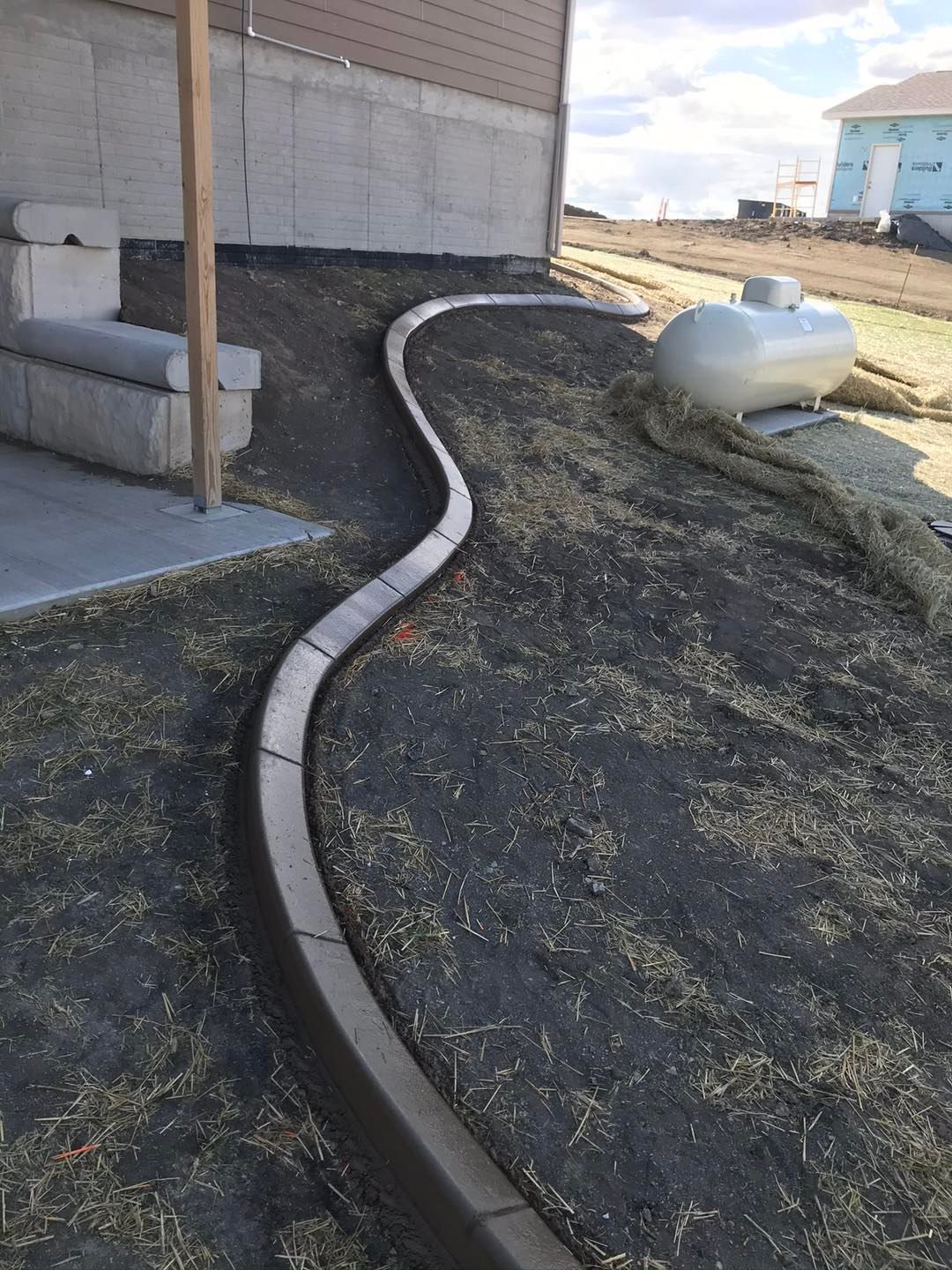 Concrete edging curves along a sloped yard next to a building and a propane tank.