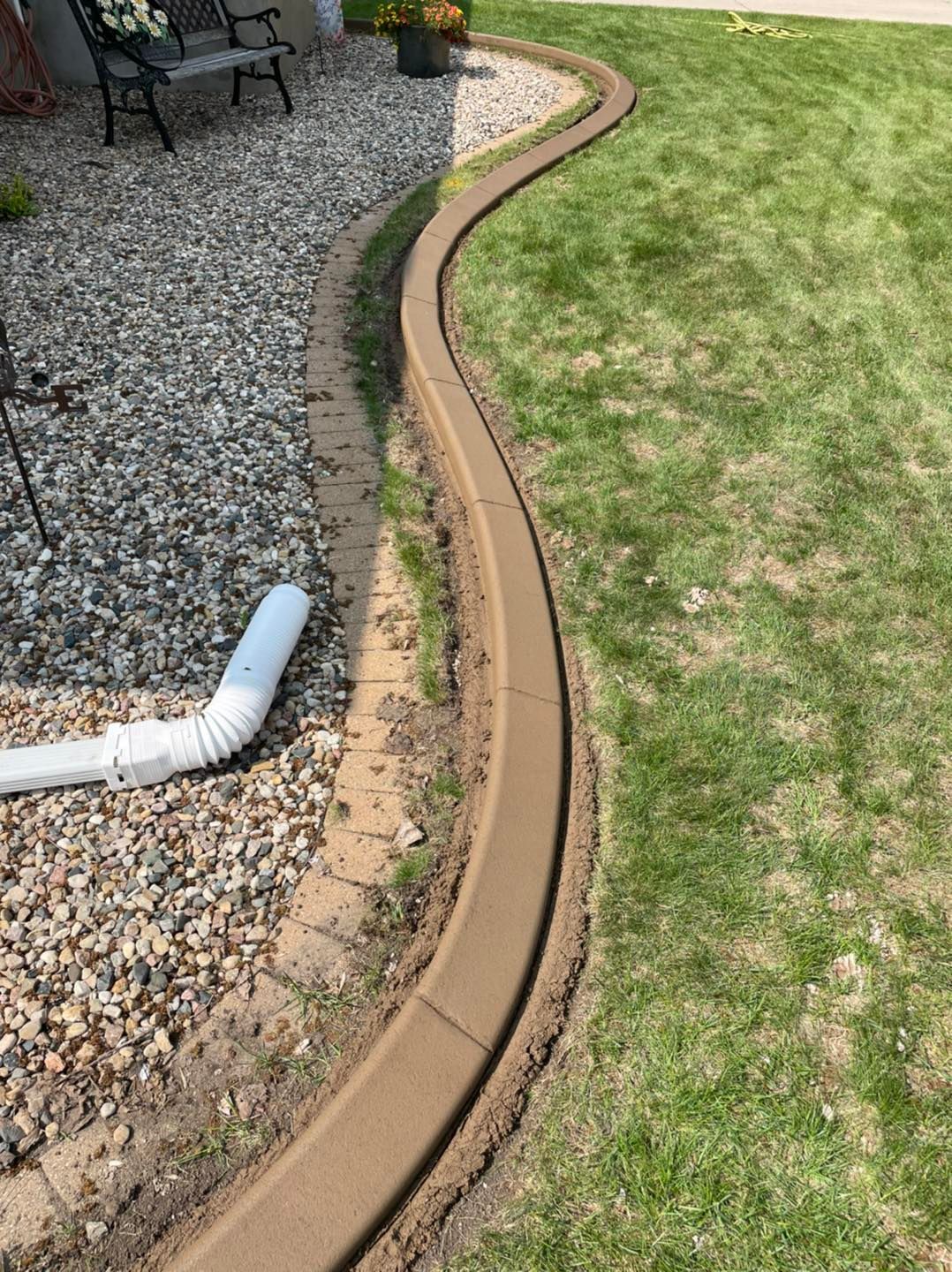 Brown concrete curbing separates a gravel area from a green lawn. White downspout is visible on the gravel.