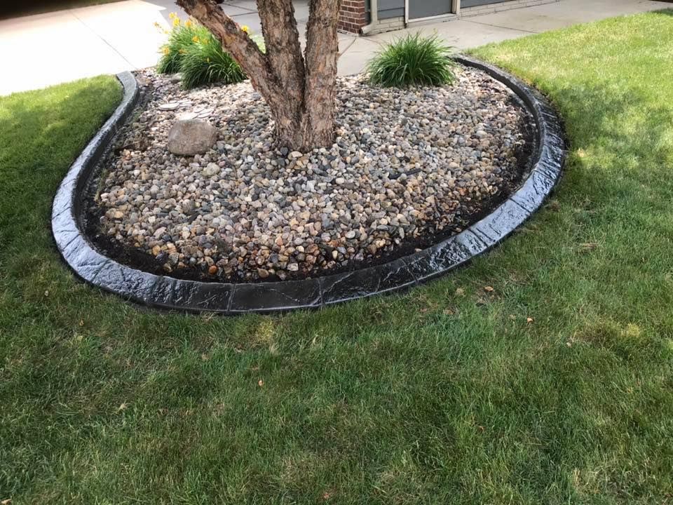 Black stamped concrete edging surrounds a tree and gravel bed in a grassy yard.