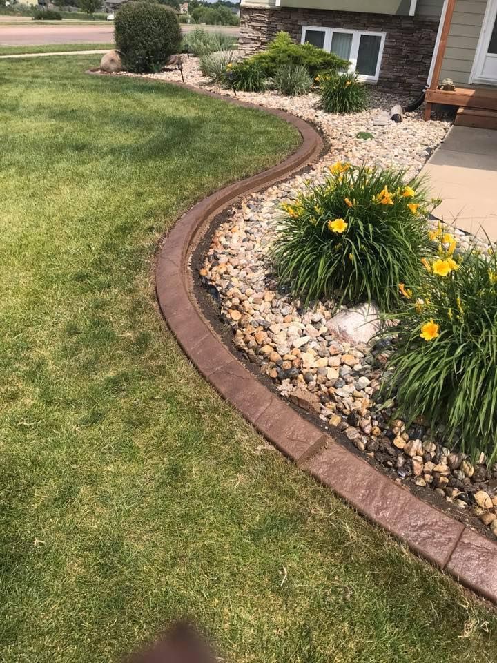Brown concrete curbing separates lawn from a rock-filled garden bed with yellow flowers and green plants.