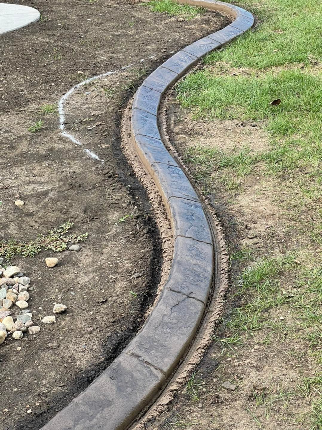 Curved concrete curbing bordering a grassy area and dirt, with a decorative pattern.