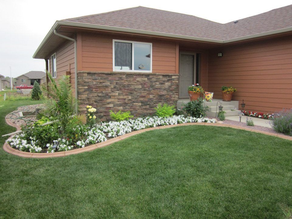 A house exterior with brown siding, a stone facade, and a curved garden bed filled with white flowers in the front yard.