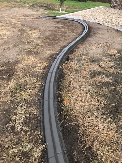 A gray concrete landscape border curves through a dirt yard, separating the soil from a gravel area in the background.