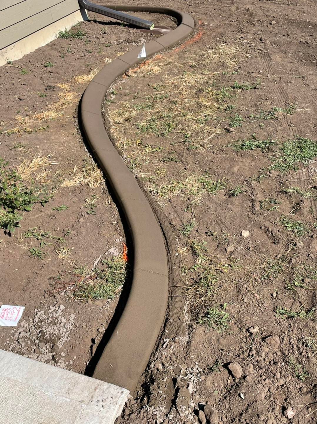 A newly installed, curved concrete landscape border runs across a patch of bare soil next to a house foundation.