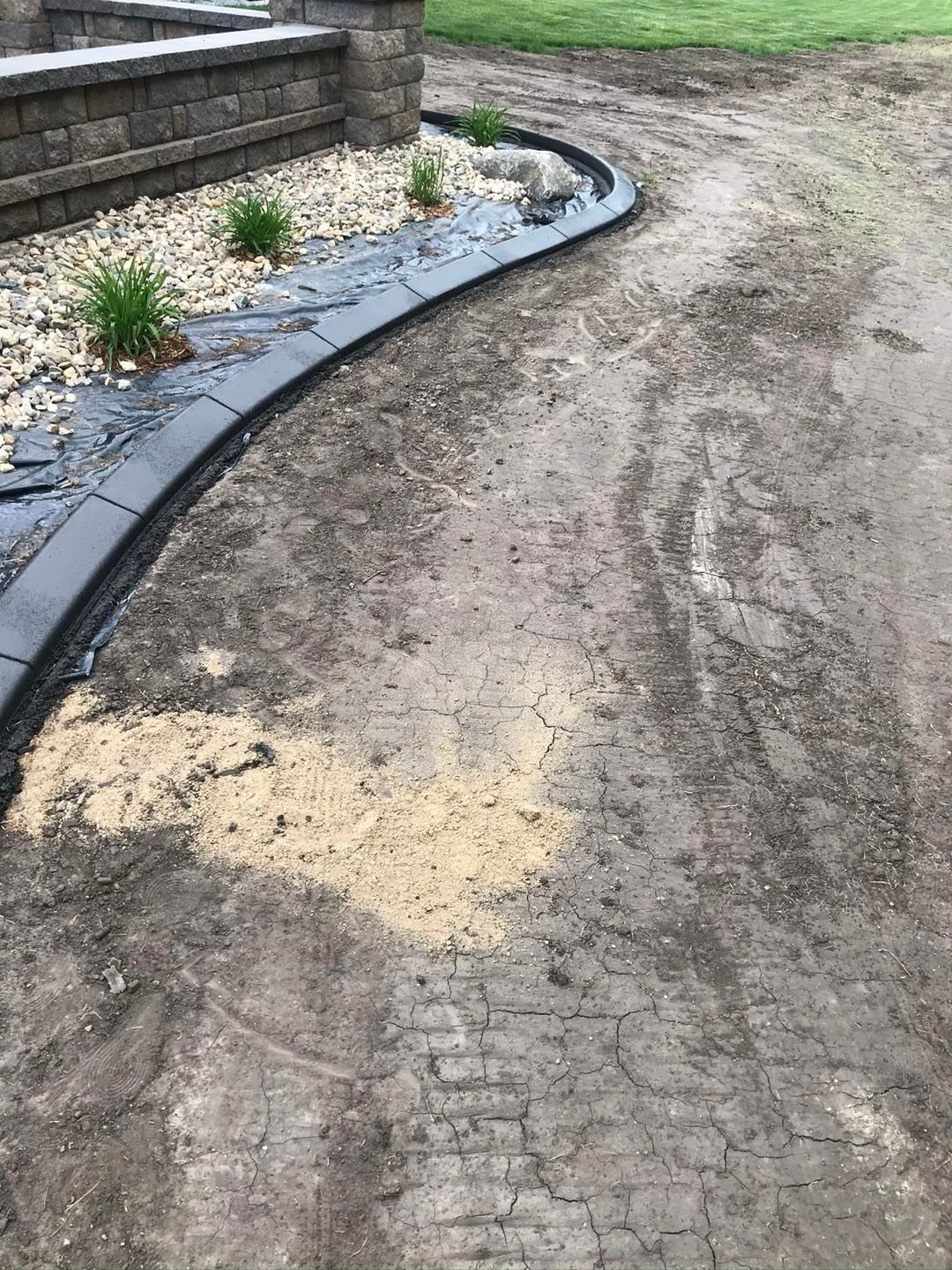 A stone retaining wall with a curved concrete border separating a landscaped gravel garden from a dirt yard.