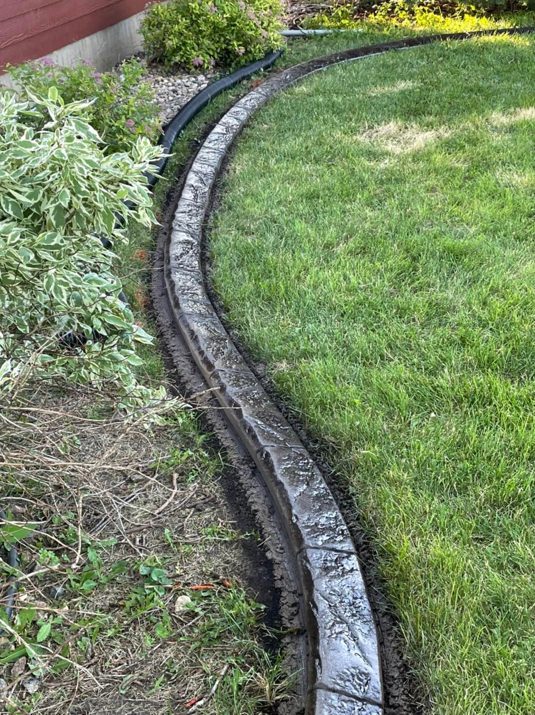 A curved, gray stone landscape border separates a grassy lawn from a mulched garden bed next to a building.