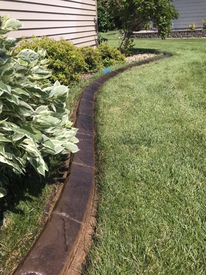 A curved, brown concrete landscape border separates a garden bed from a grassy lawn next to the side of a house.