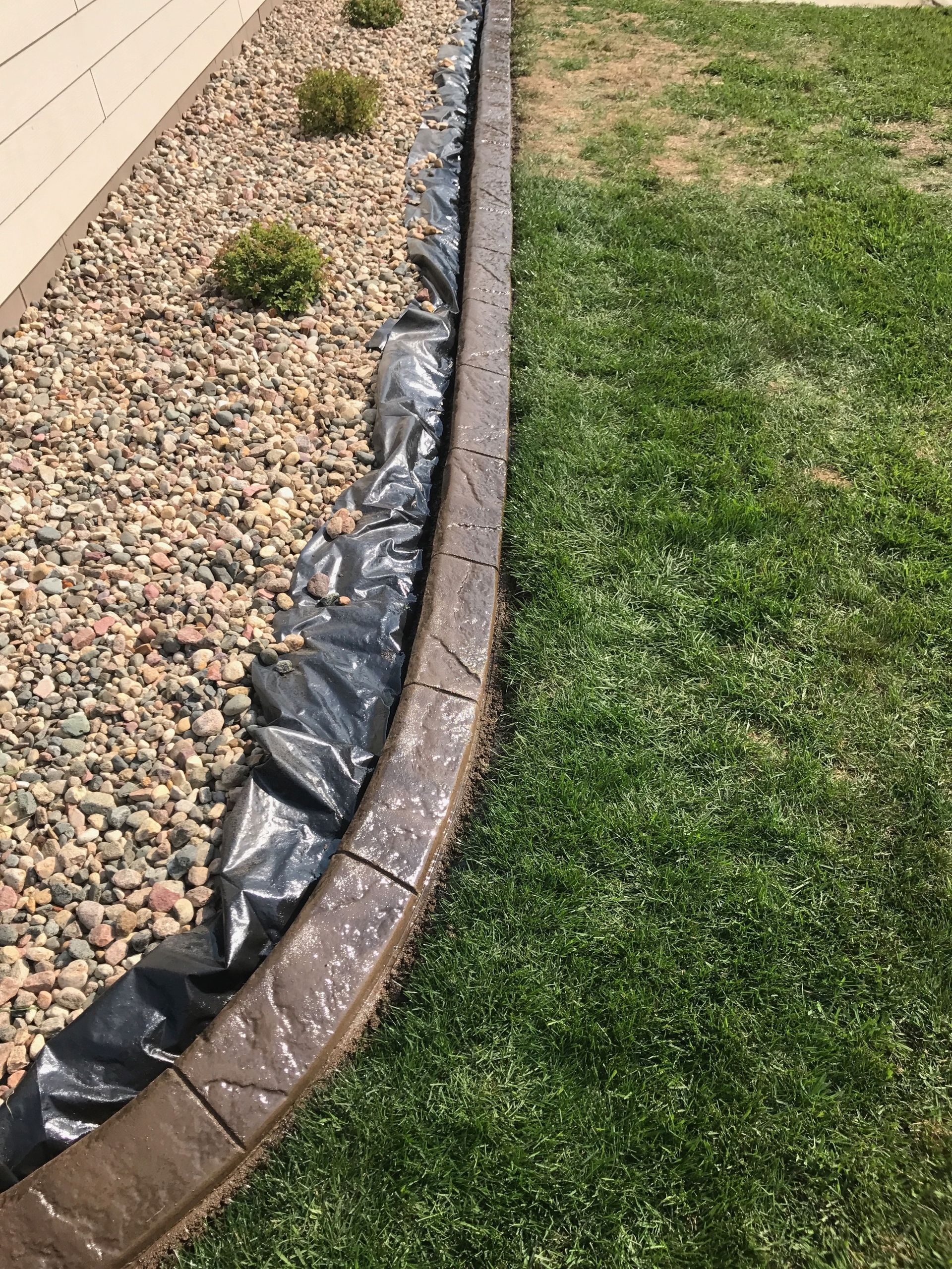 A landscape border with tan concrete edging, black plastic weed barrier, river rock mulch, and small shrubs next to a lawn.