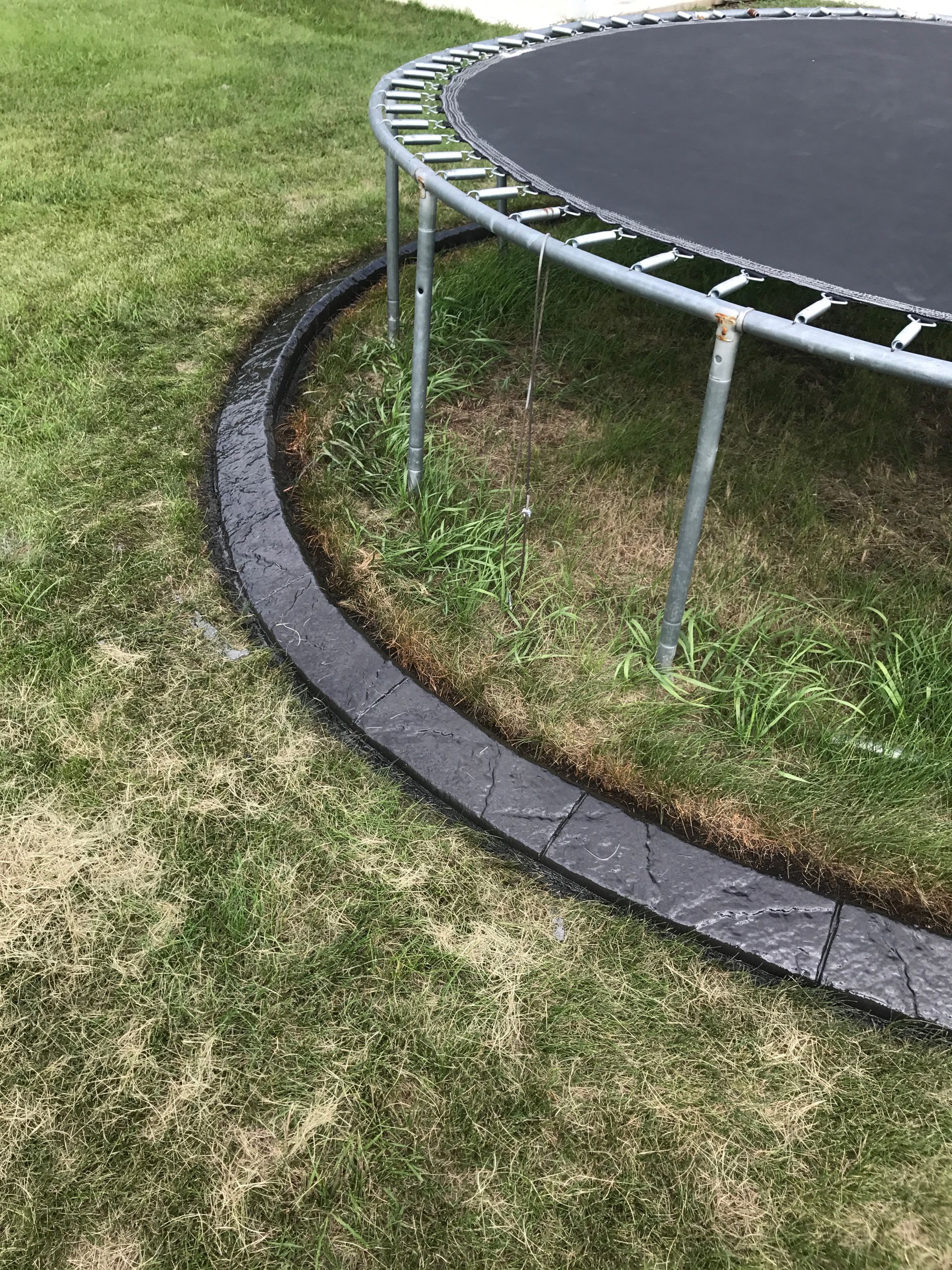 A dark gray, textured curved landscape border sits on a grass lawn next to the metal legs of a backyard trampoline.