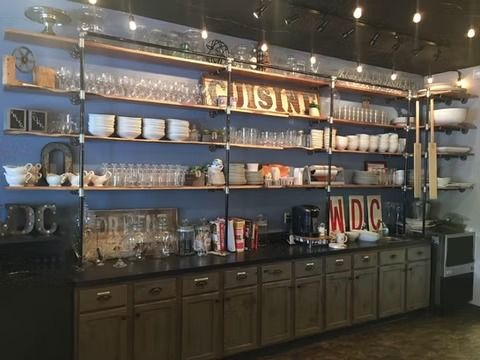 Blue restaurant wall with shelves of dishes, glassware, and decor above dark cabinets and string lights
