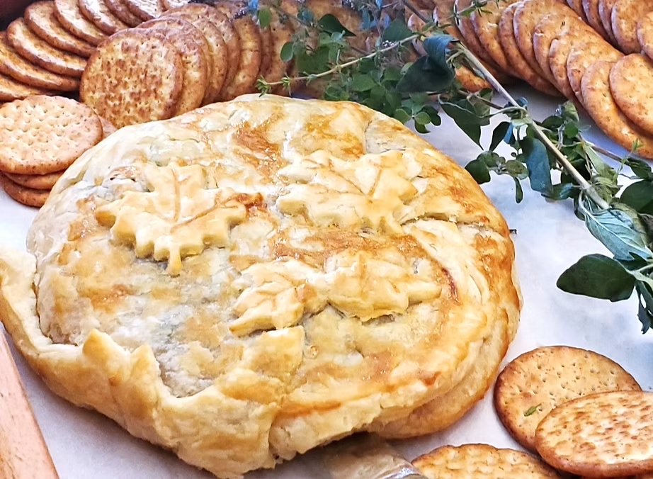 Round baked cheese pie on a plate, surrounded by crackers, sliced bread, and greenery