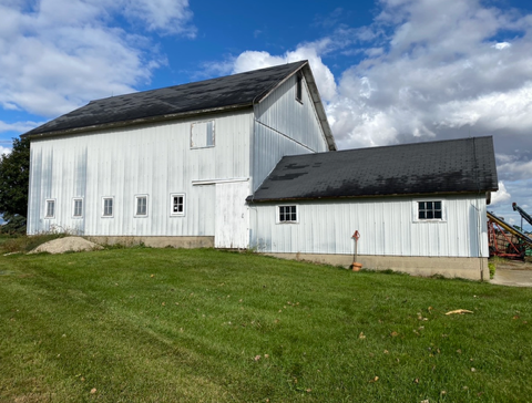 White barn and shed with dark roofs on a grassy hill under a partly cloudy blue sky