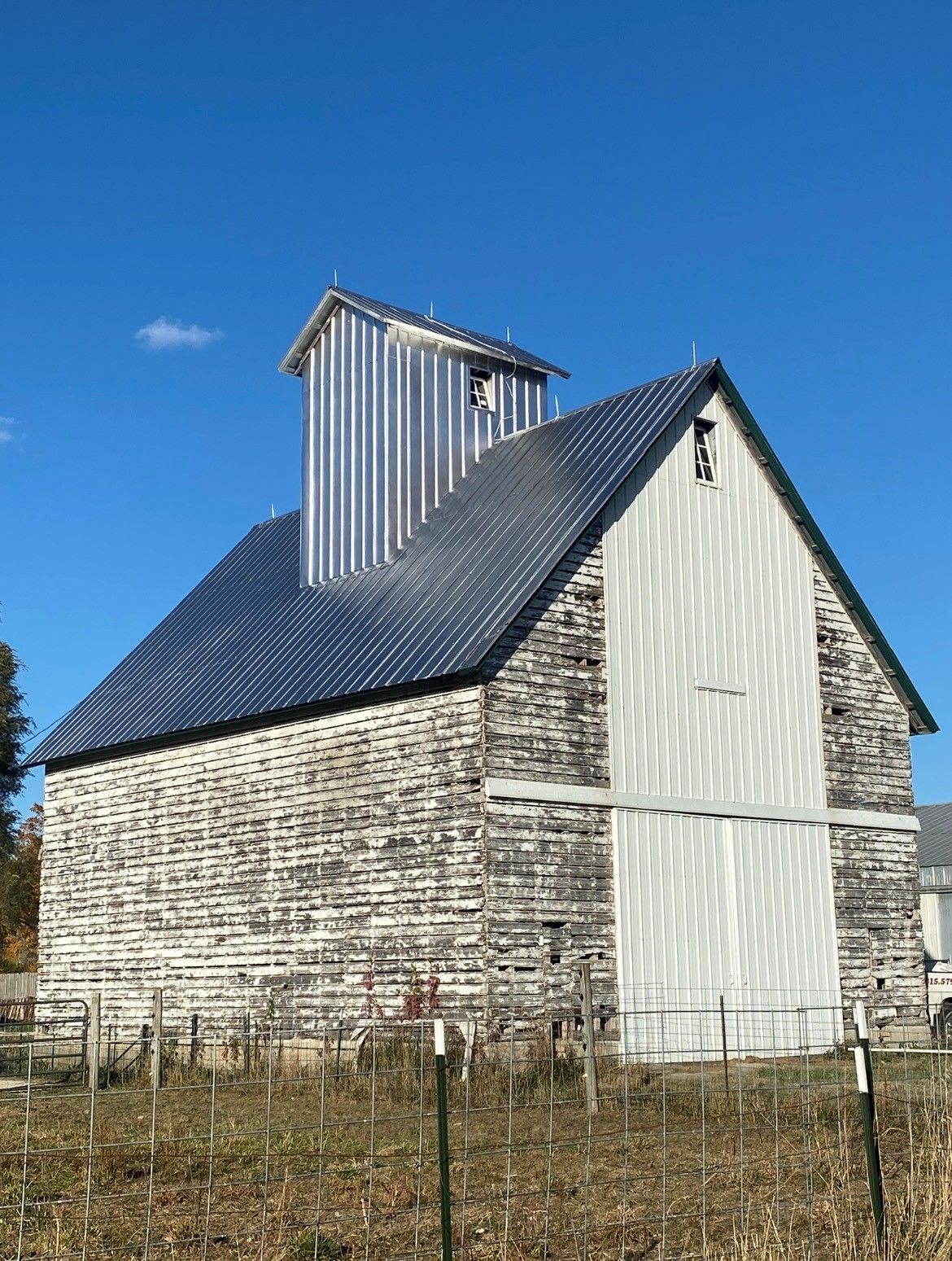 White wooden barn with a dark metal roof and cupola in a rural field under a blue sky