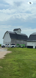 White barn with dark roof and white car on grassy field under a cloudy sky