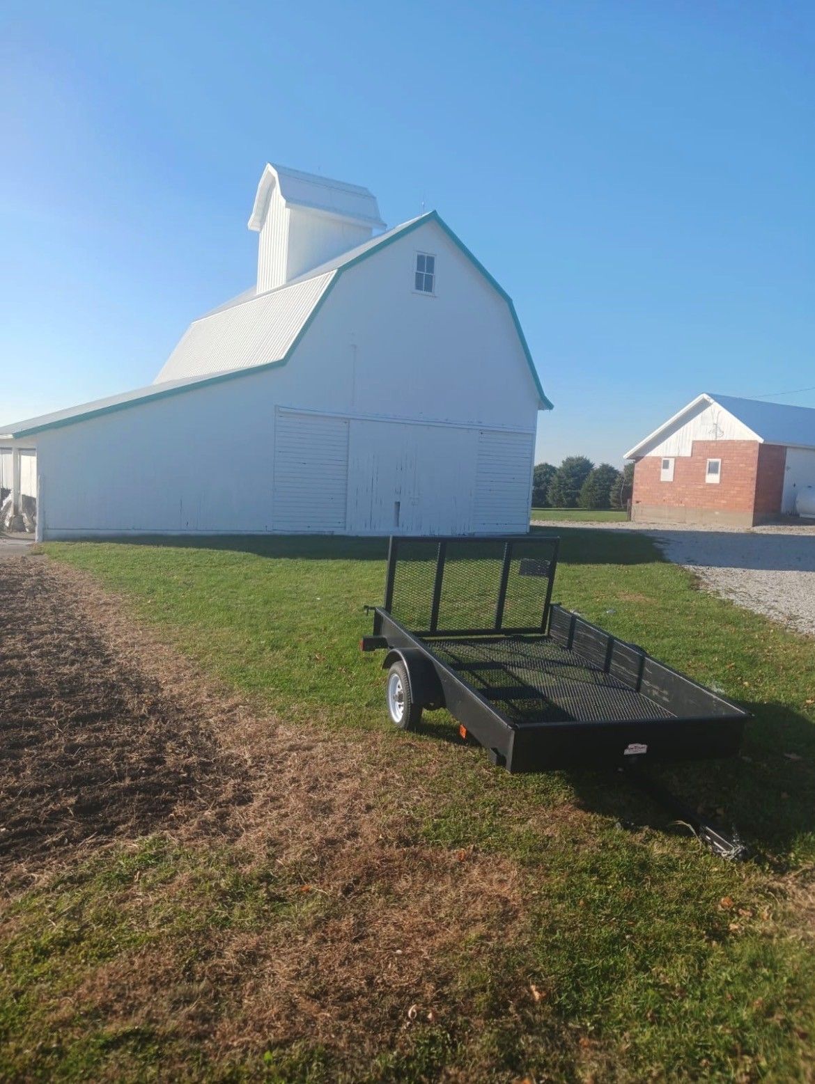 White barn on green grass with black flatbed trailer in foreground and red barn in the distance