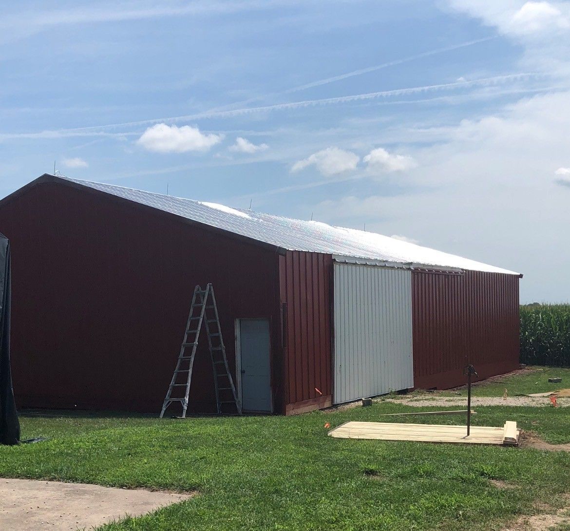 Red metal barn with white roof panels, ladder by the door, on a grassy field under a blue sky.