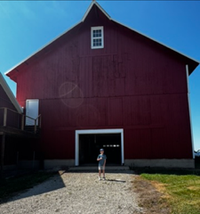 Large red barn with a person standing in the open doorway on a gravel path under a blue sky