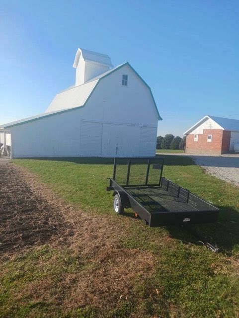 White barn beside a grassy path with a black utility trailer in the foreground and a brick house in the background
