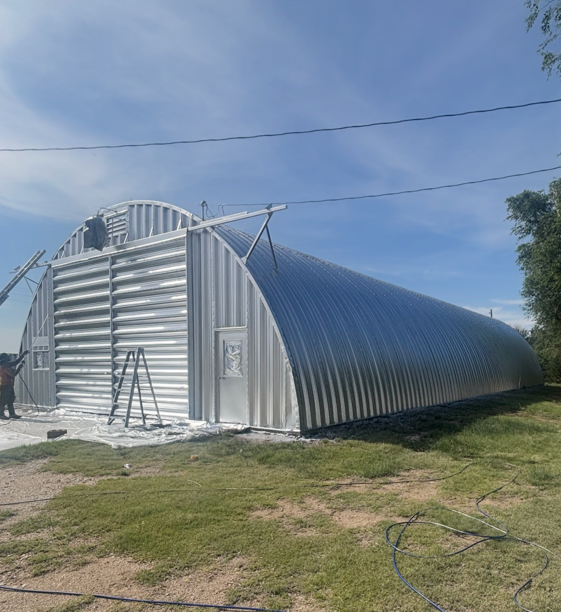 Silver corrugated metal Quonset-style building under construction on a grassy lot under a blue sky