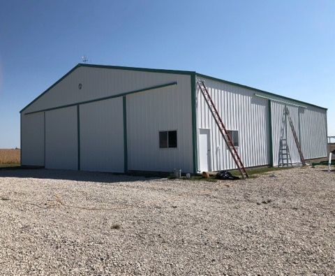 Large white metal storage building with a green roof and red ladder on a gravel lot under a blue sky