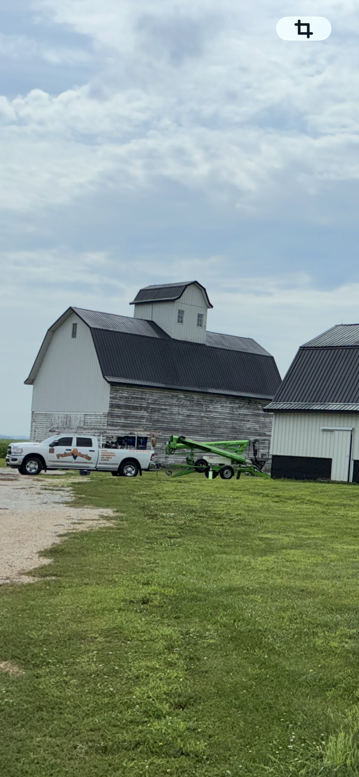 Weathered barn with a damaged roof and parked white trucks under a cloudy sky on a grassy lot