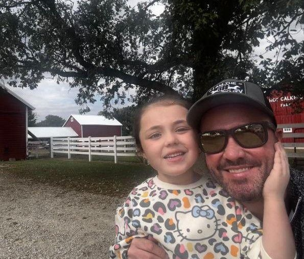 Man and child smile in front of a farm with red barns, a white fence, and a large tree.