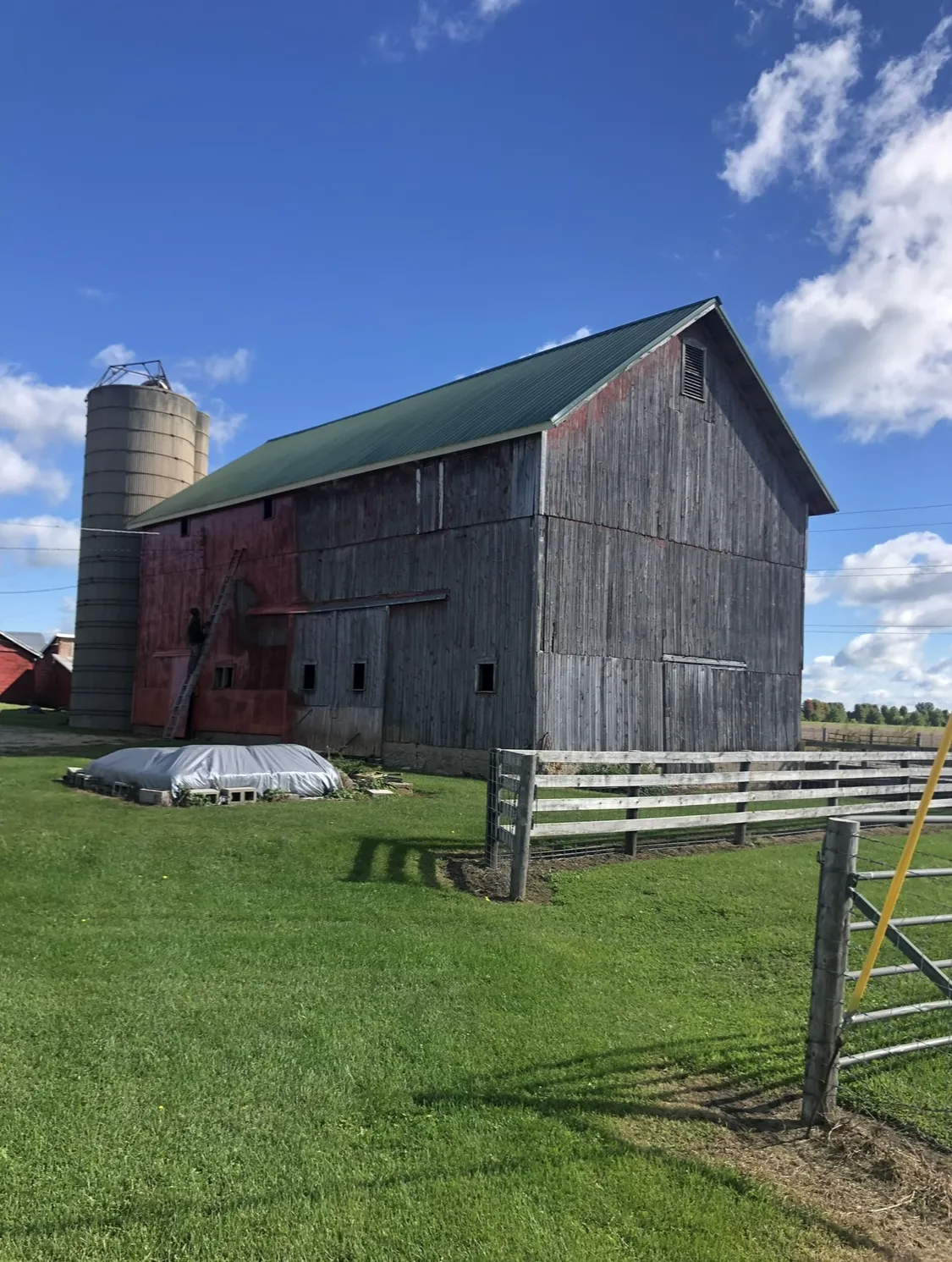 Weathered red and gray barn with a silo on a green field under a blue sky with white clouds.