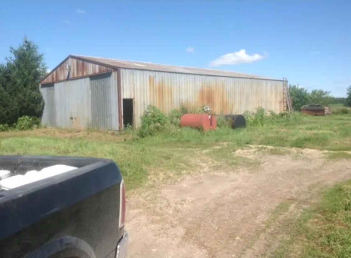 Rusty metal barn in a field with tall grass, an open door, and a truck in the foreground.