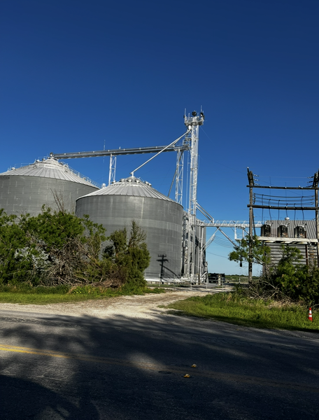 Large metal grain silos and conveyor system against a blue sky, beside a road.