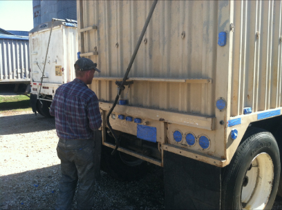 Man inspecting the back of a beige truck. Another truck in the background. Sunny outdoor setting.