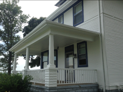 White two-story house with a covered porch supported by columns. The porch has a white railing. Blue trim on windows.