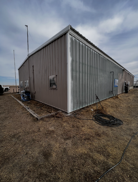 Metal building with gray siding in a grassy field under a cloudy sky.