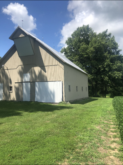 Tan barn with white doors and a large green tree under a blue sky.