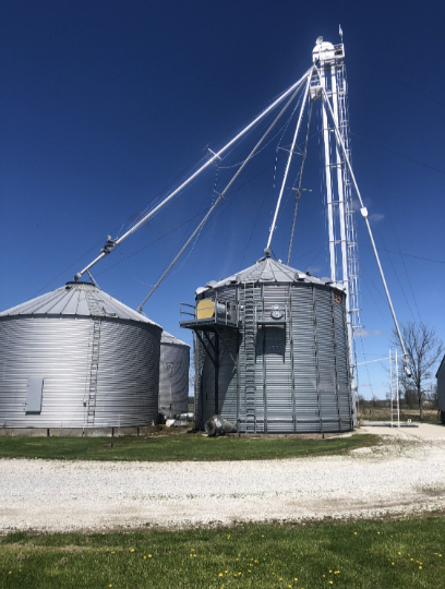 Grain silos and a tall elevator tower against a blue sky, on a gravel surface.