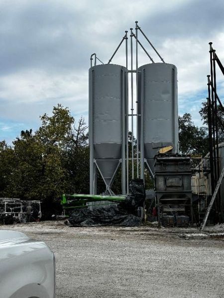 Two gray industrial silos stand outdoors against a cloudy sky.