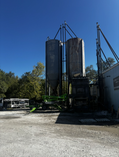 Two metal silos with support structures, a truck, and a building under a blue sky.