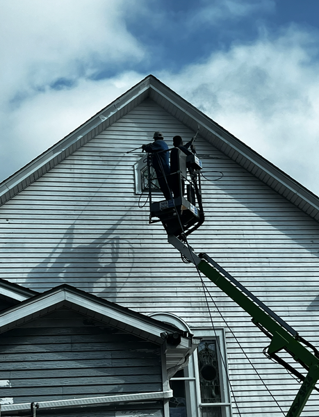 Two workers in a lift cleaning the siding of a light-colored house against a cloudy sky.