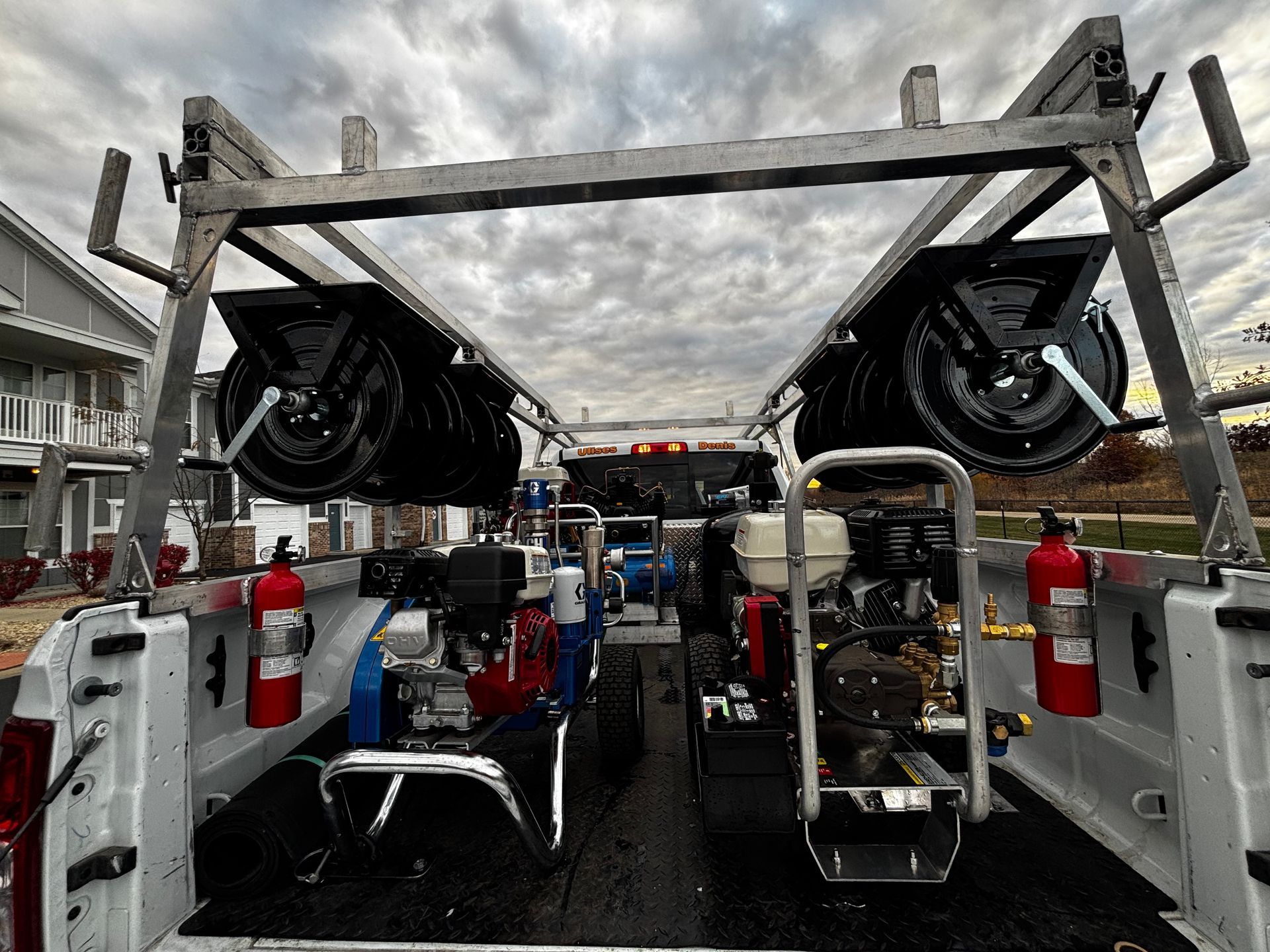 Truck bed with pressure washing equipment: hose reels, pumps, fire extinguishers, and roof rack. Overcast sky.