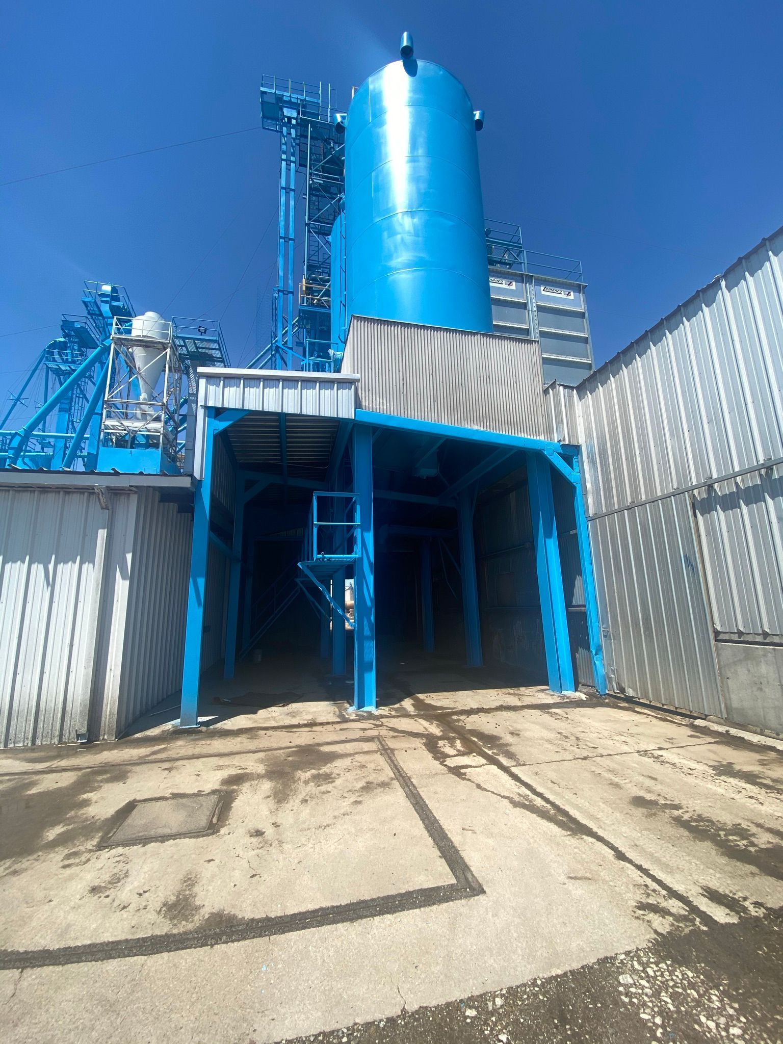 Blue industrial plant with tall silo, metal structure, and concrete ground.