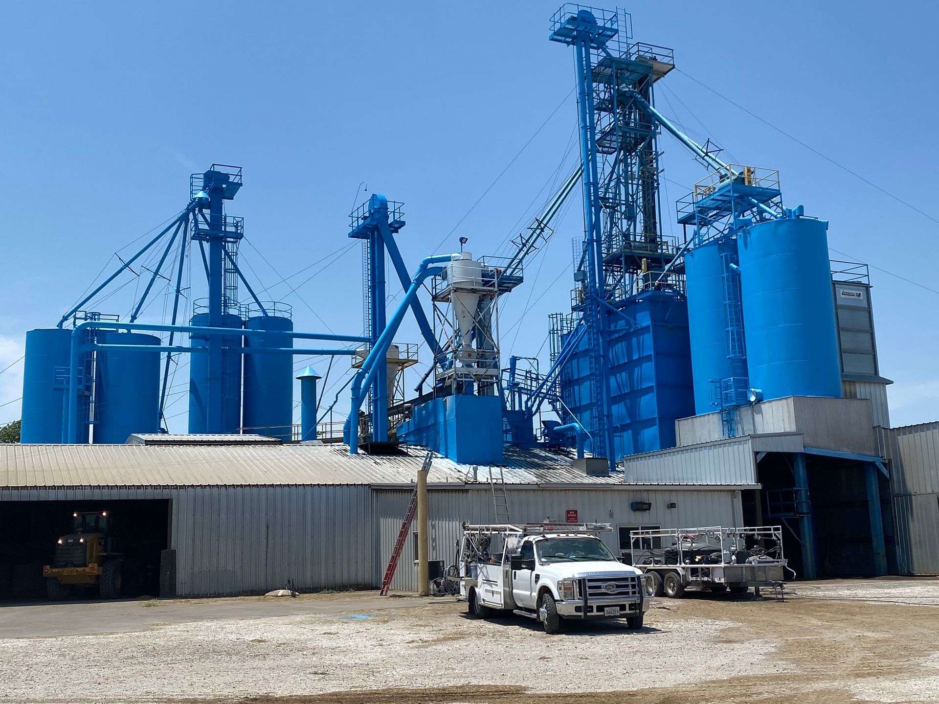 Blue industrial grain processing plant with silos and service vehicles in front.