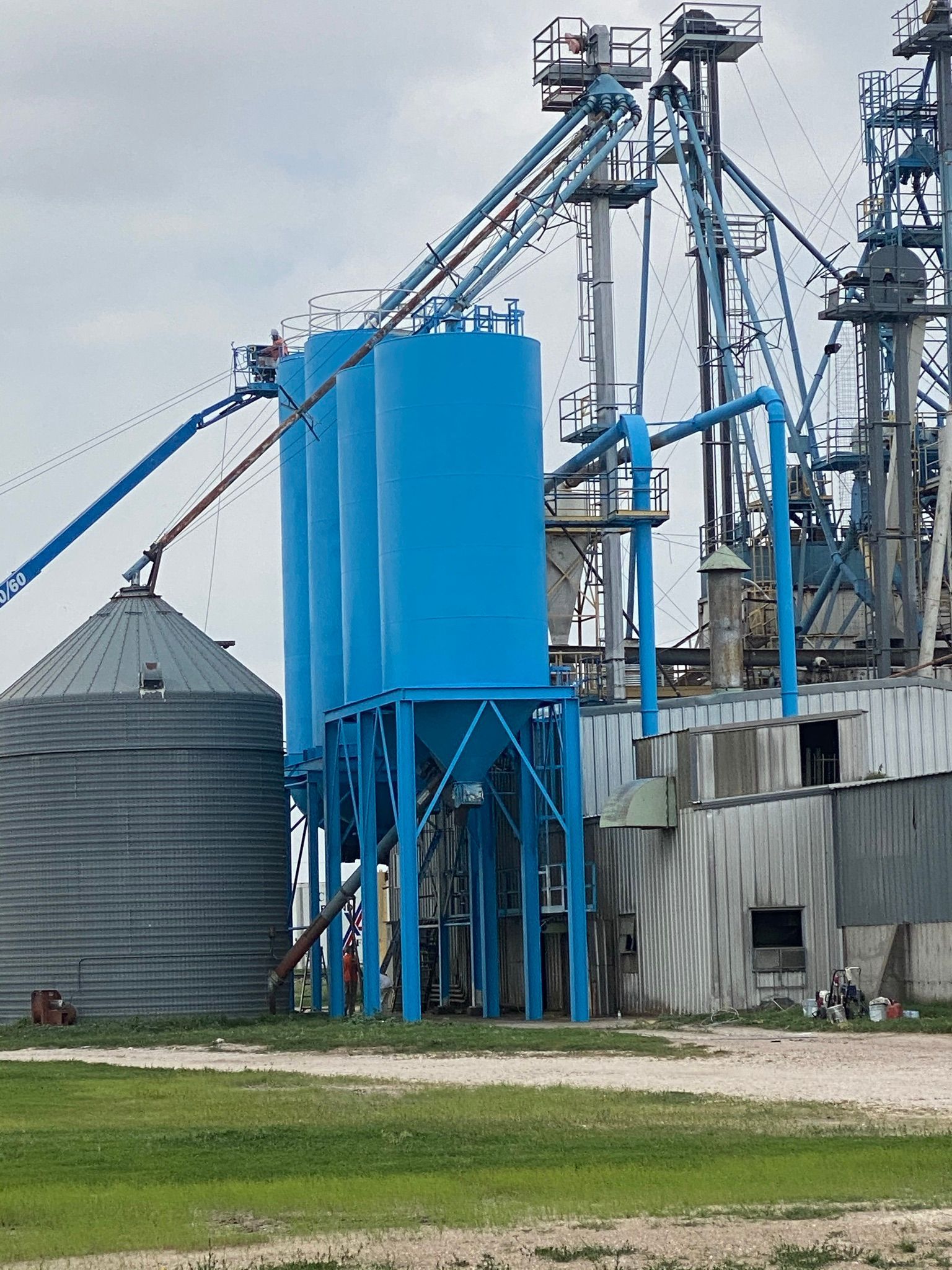 Blue silos and industrial machinery on a gray day, set on green grass.