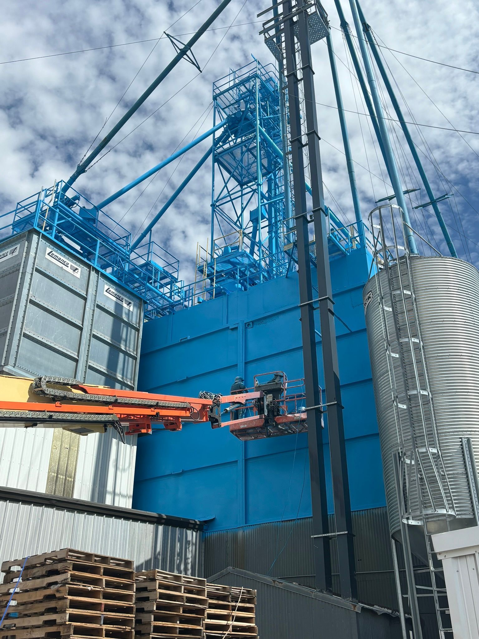 Man in lift working on blue industrial structure; sky background.