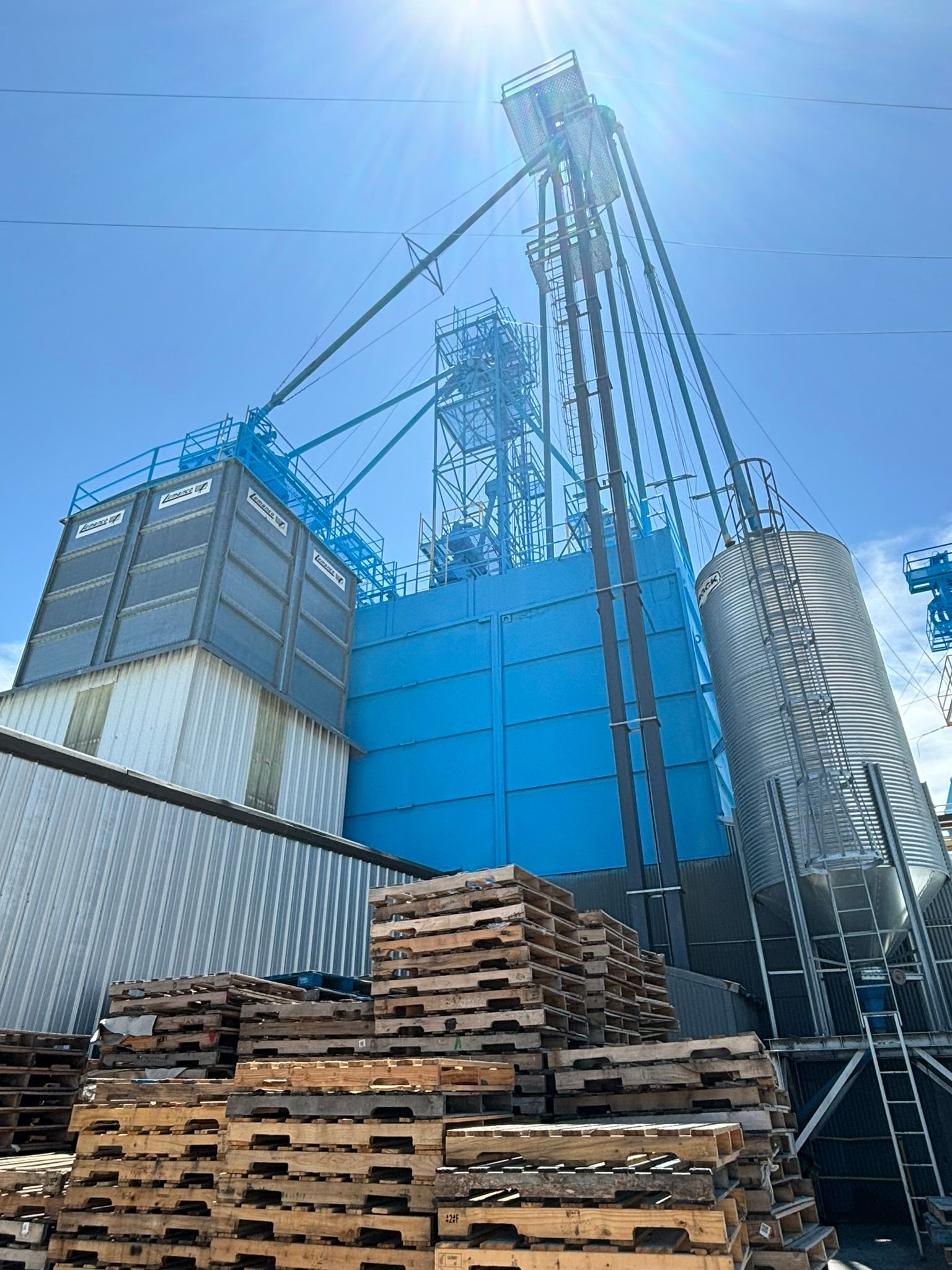Blue industrial grain elevator with tall metal structure and stacks of wooden pallets.