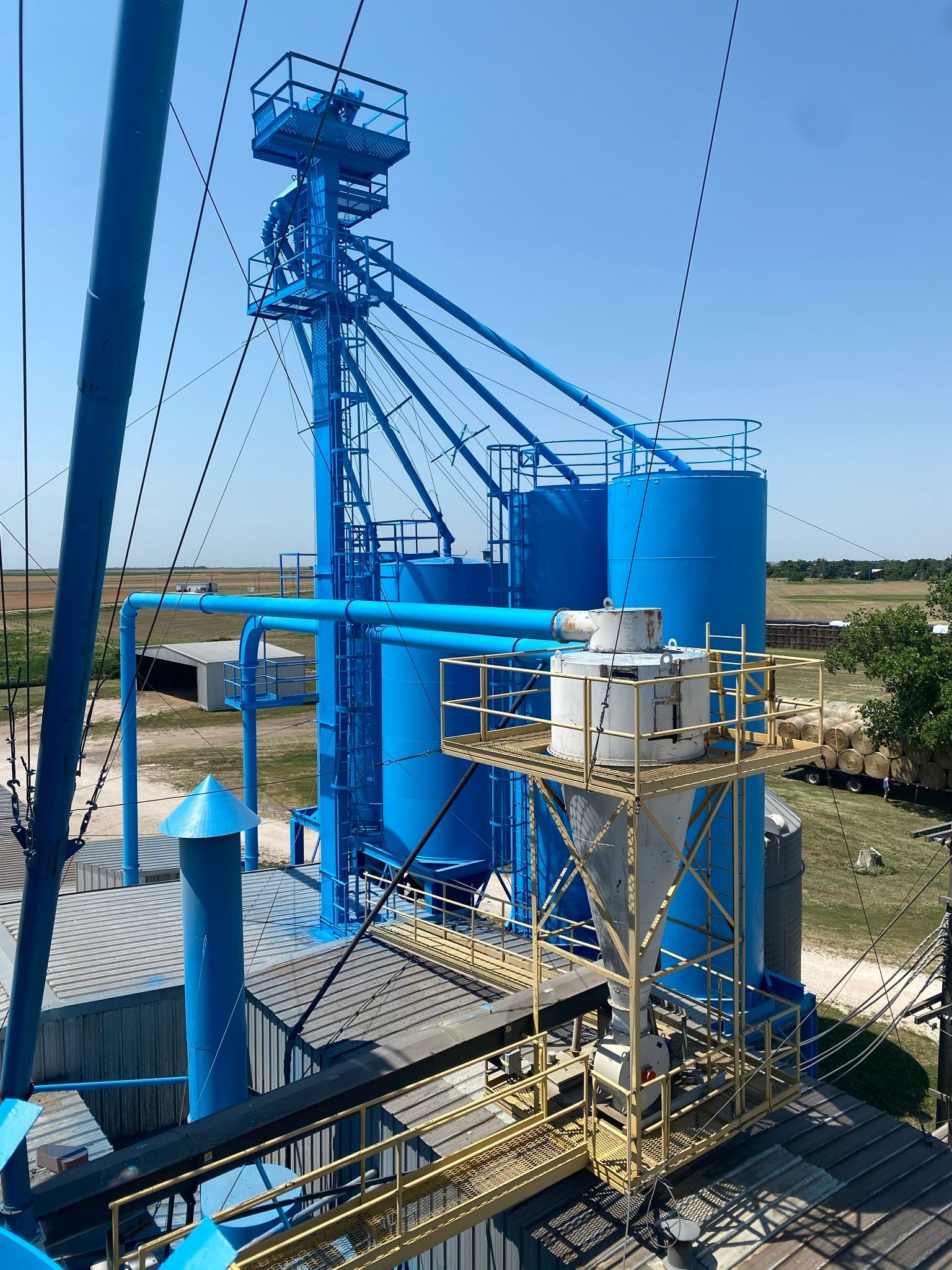 Blue industrial grain processing facility with silos, pipes, and conveyor systems against a clear sky.