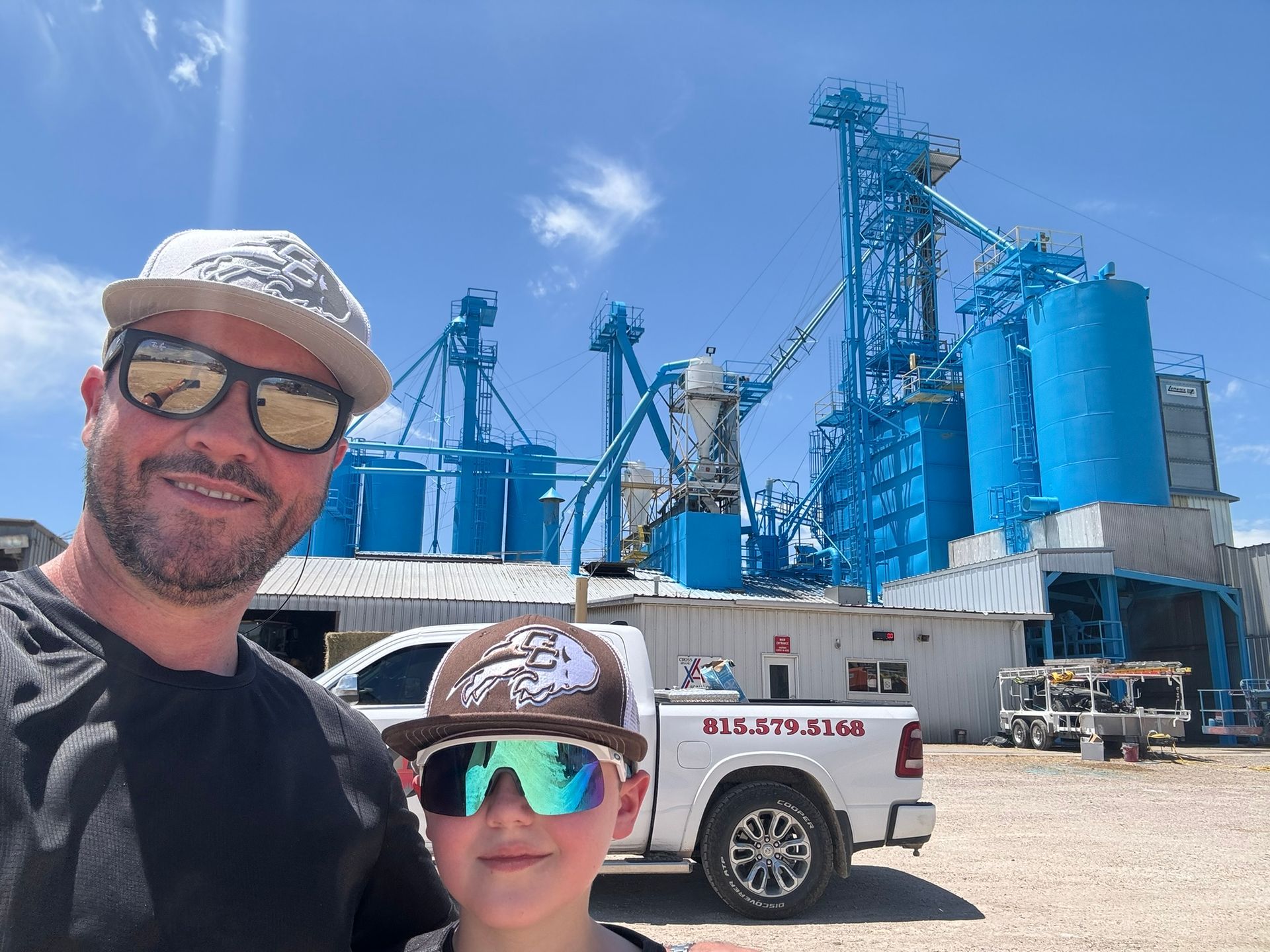 Man and boy in front of a bright blue industrial building and a white truck on a sunny day.