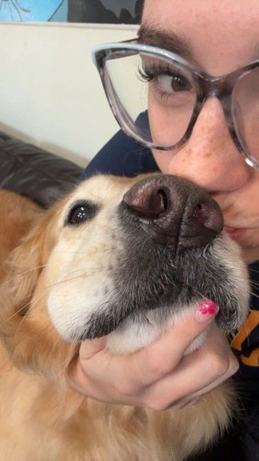 Woman kissing a golden retriever dog; both close-up, indoors. Dog has dark nose and mouth. Woman wears glasses.