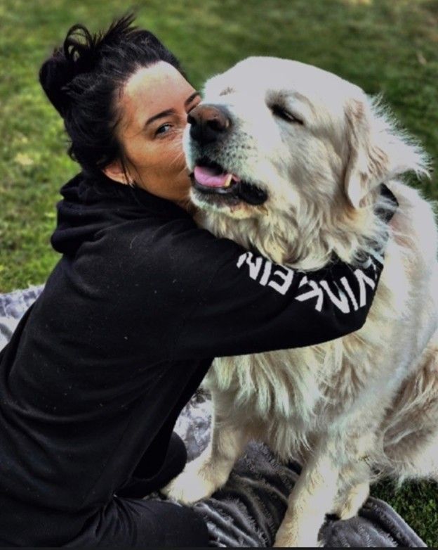 Woman in black hoodie kissing a large, cream-colored dog on the cheek outdoors, dog panting.