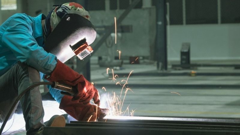 Welder wearing a helmet and gloves welding metal, creating sparks in a workshop.