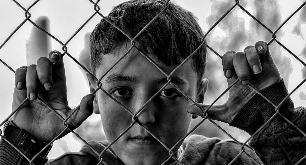 A black and white photo of a boy behind a chain link fence